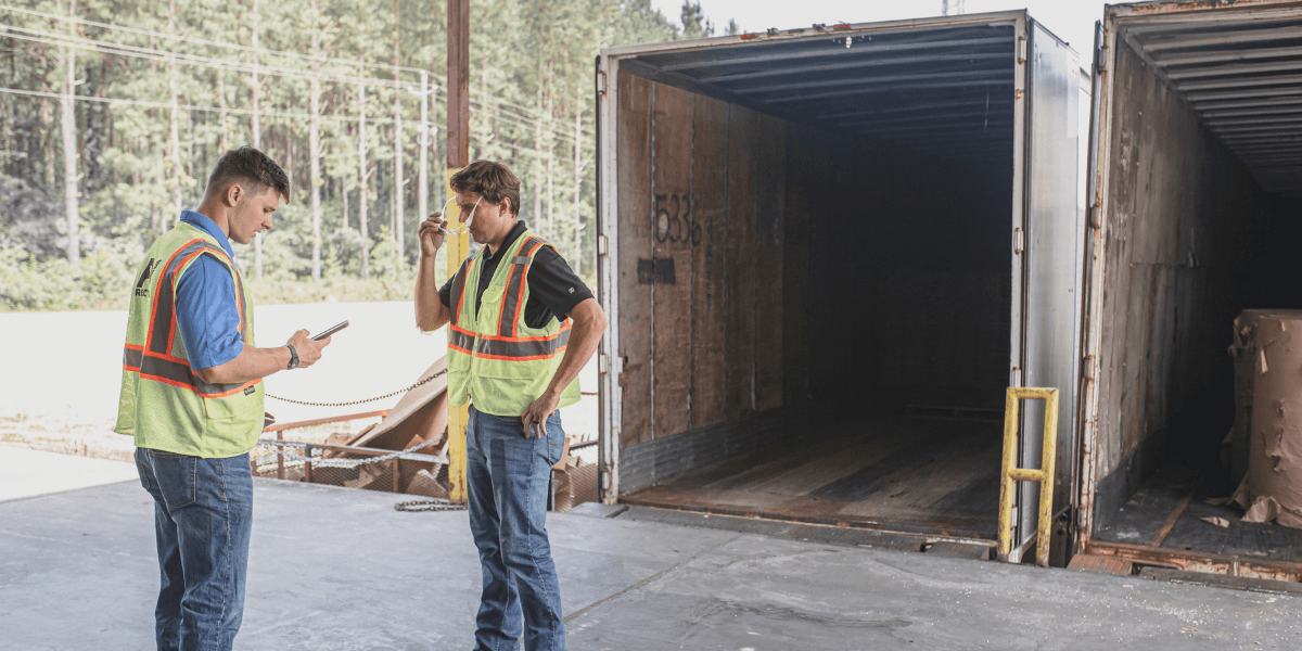Two FV Recycling team members in safety vests review a drop-trailer pickup at a loading dock beside open trailers
