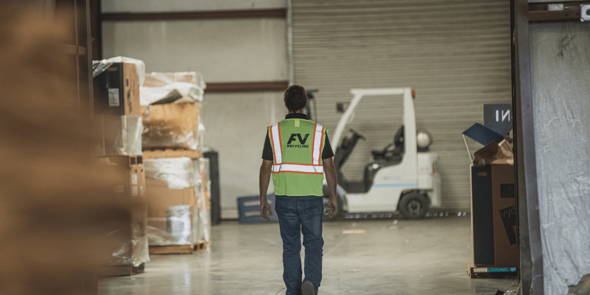 FV Recycling worker in a safety vest walks through a recycling facility past stacked cardboard and a forklift.