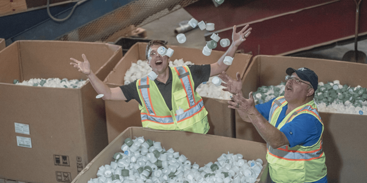 Two recycling workers in safety vests toss plastic bottle caps above gaylord boxes of sorted rigid plastic for recycling