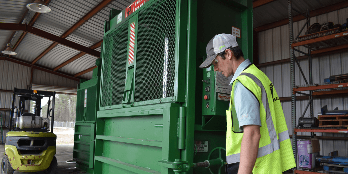 Technician inspecting industrial cardboard baler machine inside warehouse to troubleshoot baler jam and improve recycling equipment performance