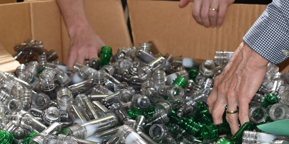 Hands sorting through a box of clear and green PET plastic bottle preforms to check for contamination