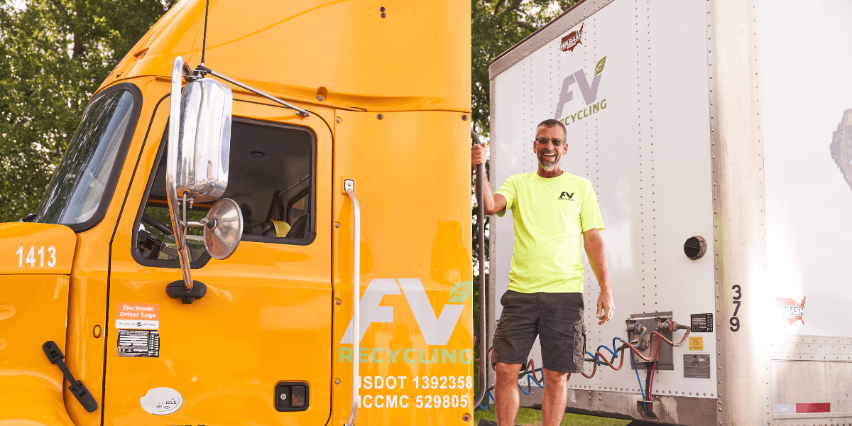 FV Recycling driver standing between a yellow truck and trailer at a commercial recycling pickup site.