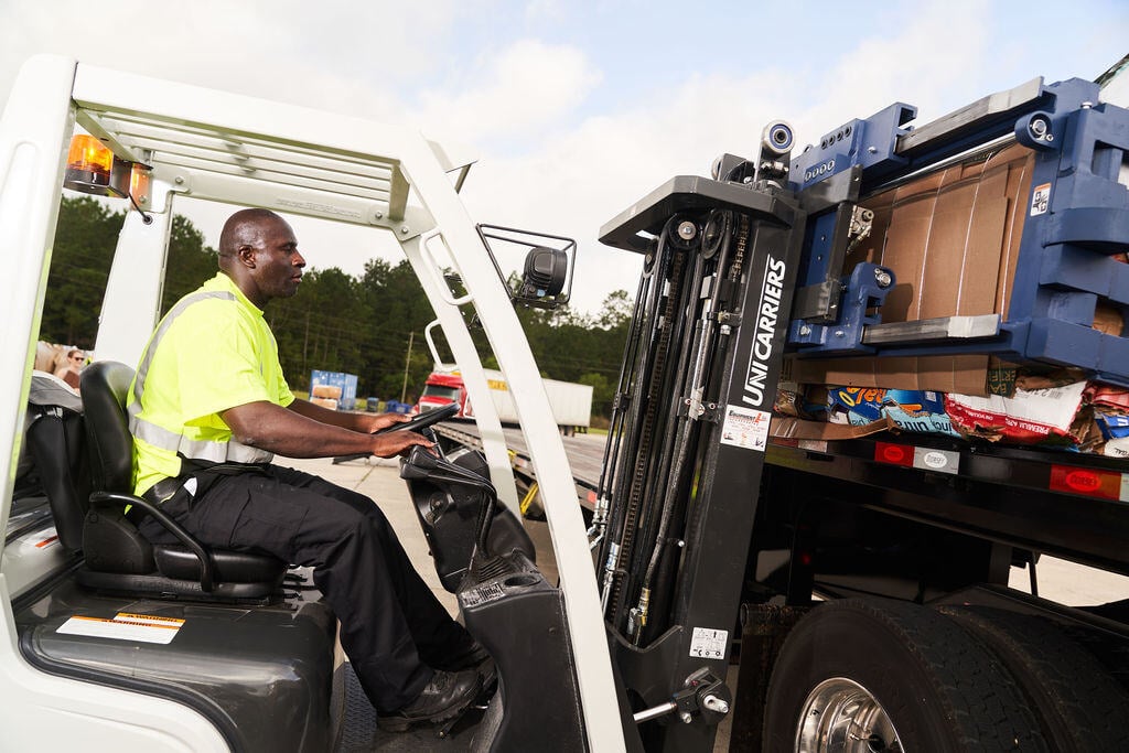 FV Recycling worker in a safety shirt driving a fork lift, lifting up a large blue recycling dumpster