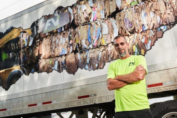 Male FV Recycling truck driver with a highlighter yellow safety shirt standing in front of  the side of an 18-wheeler trailer with a recycling graphic